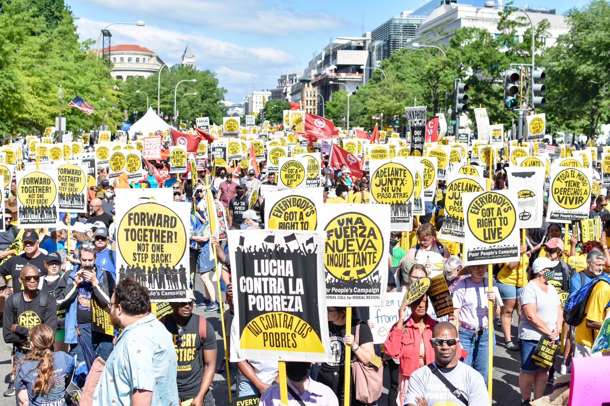 Photo of crowd filling the street on June 18, many holding Poor People's Campaign signs in yellow, black and white with slogans like Forward Together, Lucha Contra La Pobreza, Everybody Has the Right to Live.